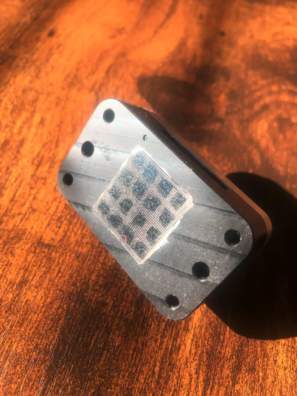 Machined aluminium chip carrier with four screw holes holding a square silicon chip whose 4 x 4 grid of windows (the molecular aquarium array) catches the light.