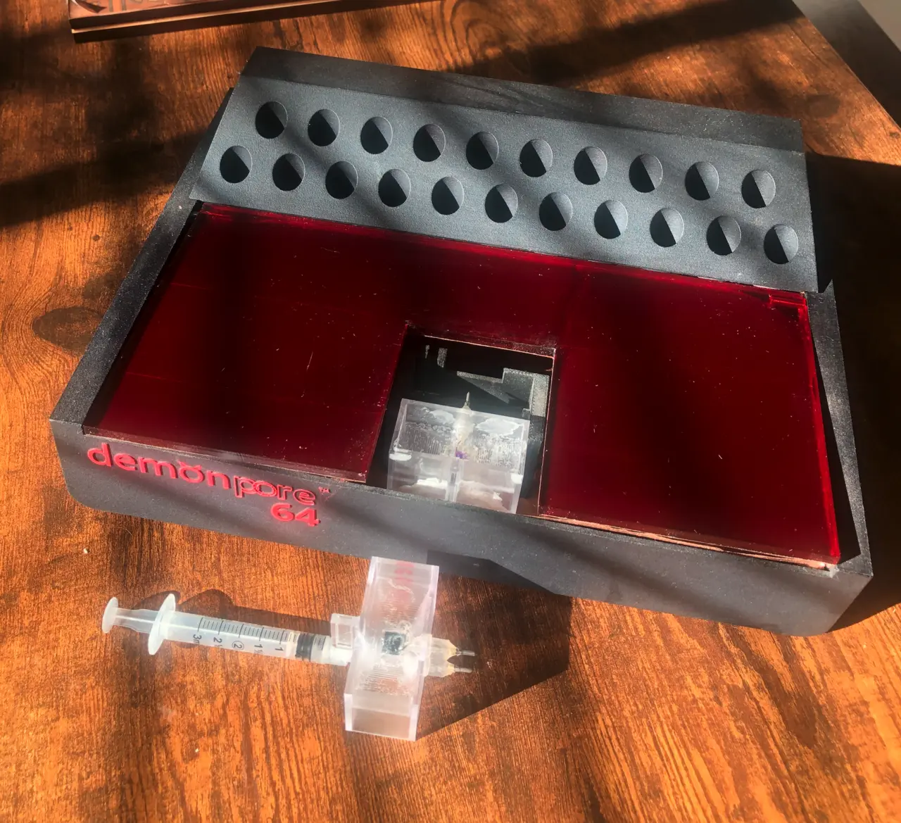 A matte black tray with two rows of teardrop-shaped wells, a red translucent inset holding a clear flow cell, and a syringe in front